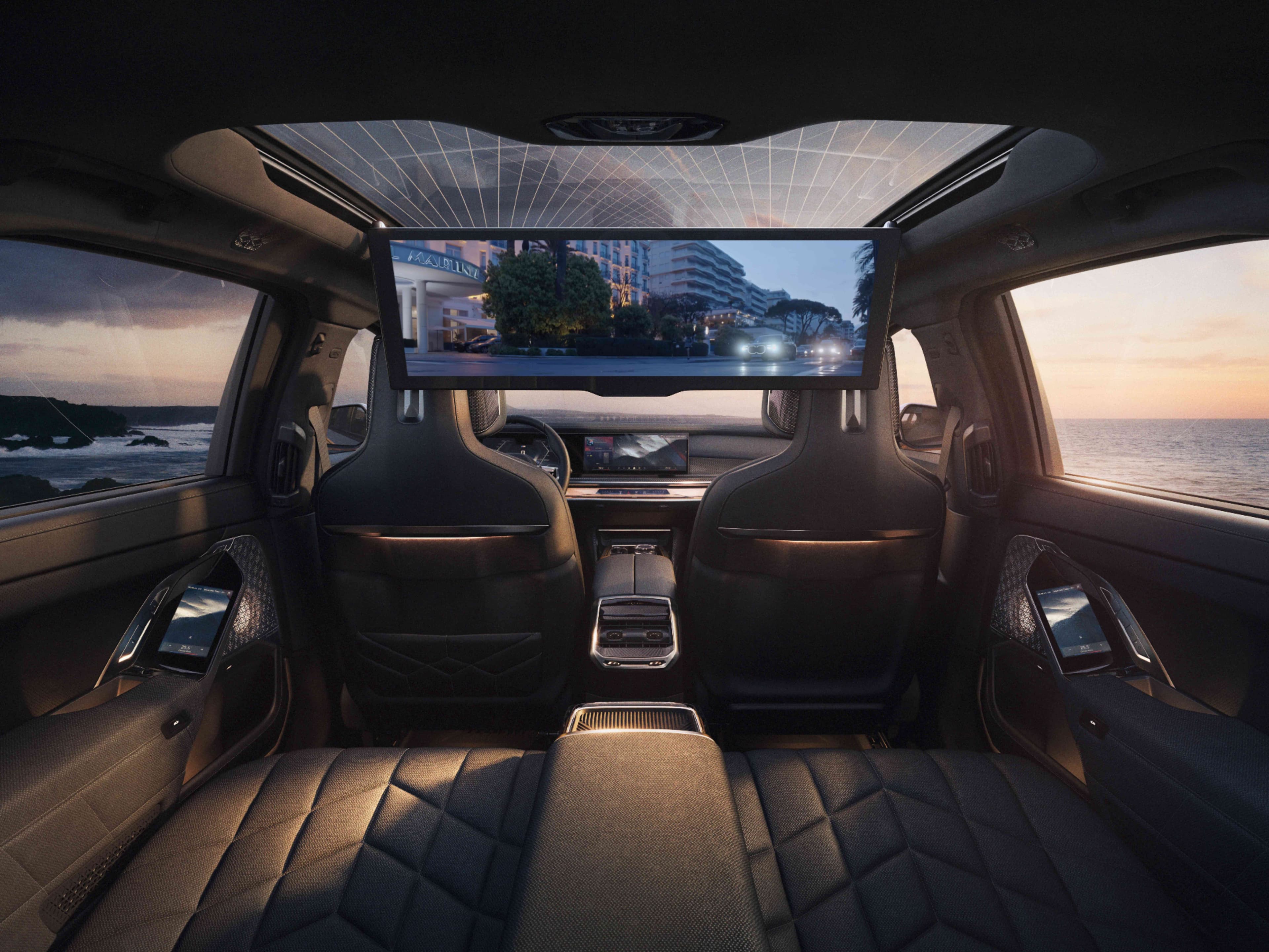 Interior view of a luxury car's back seat with a large screen displaying a cityscape at sunset.
