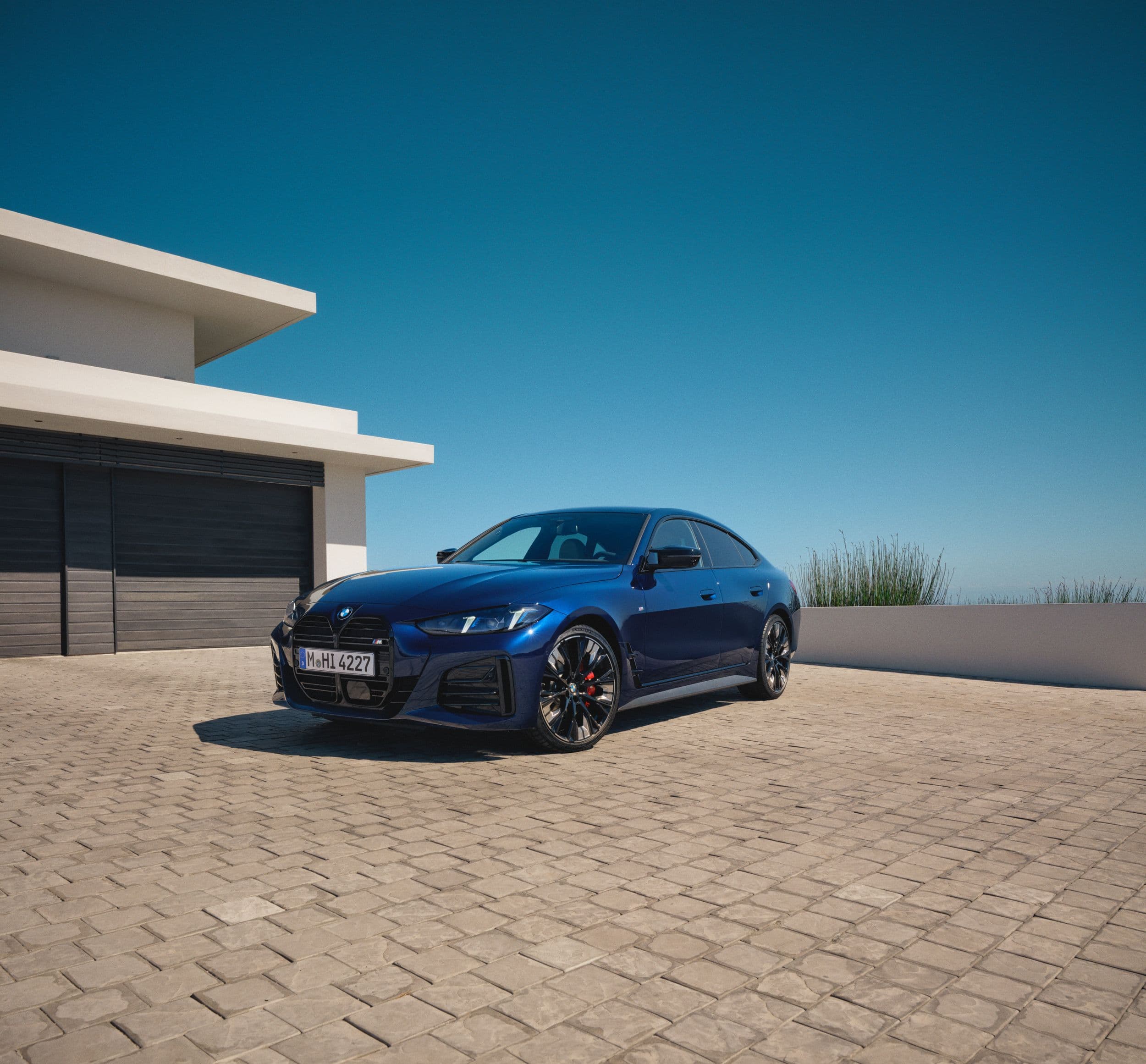A sleek, dark blue BMW M4 Coupe is parked on a paved driveway in front of a modern house.