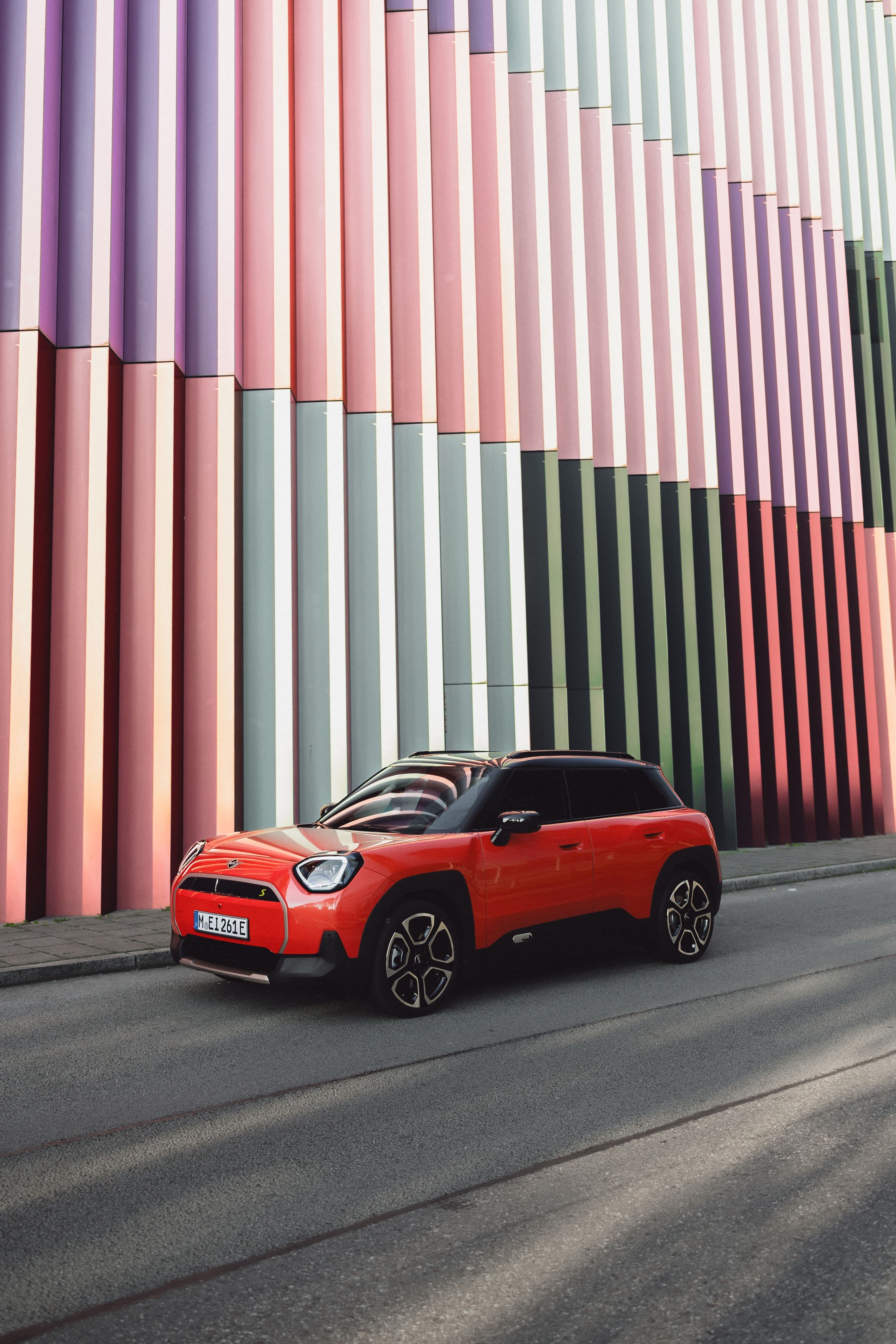 A bright orange Mini Cooper Countryman parked on a street in front of a building with colorful vertical columns.