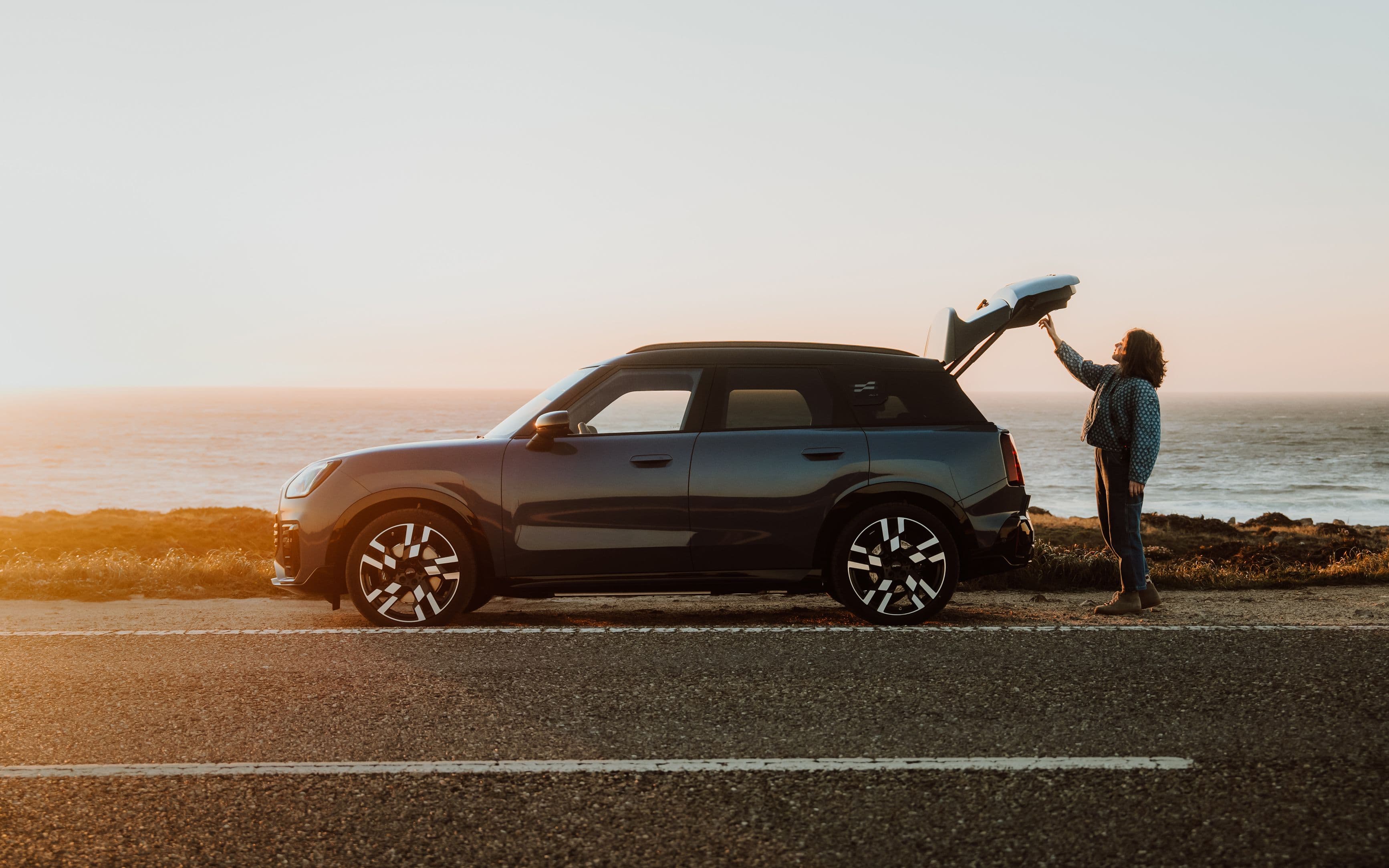 Woman loading luggage into the open trunk of a dark SUV parked by the ocean.