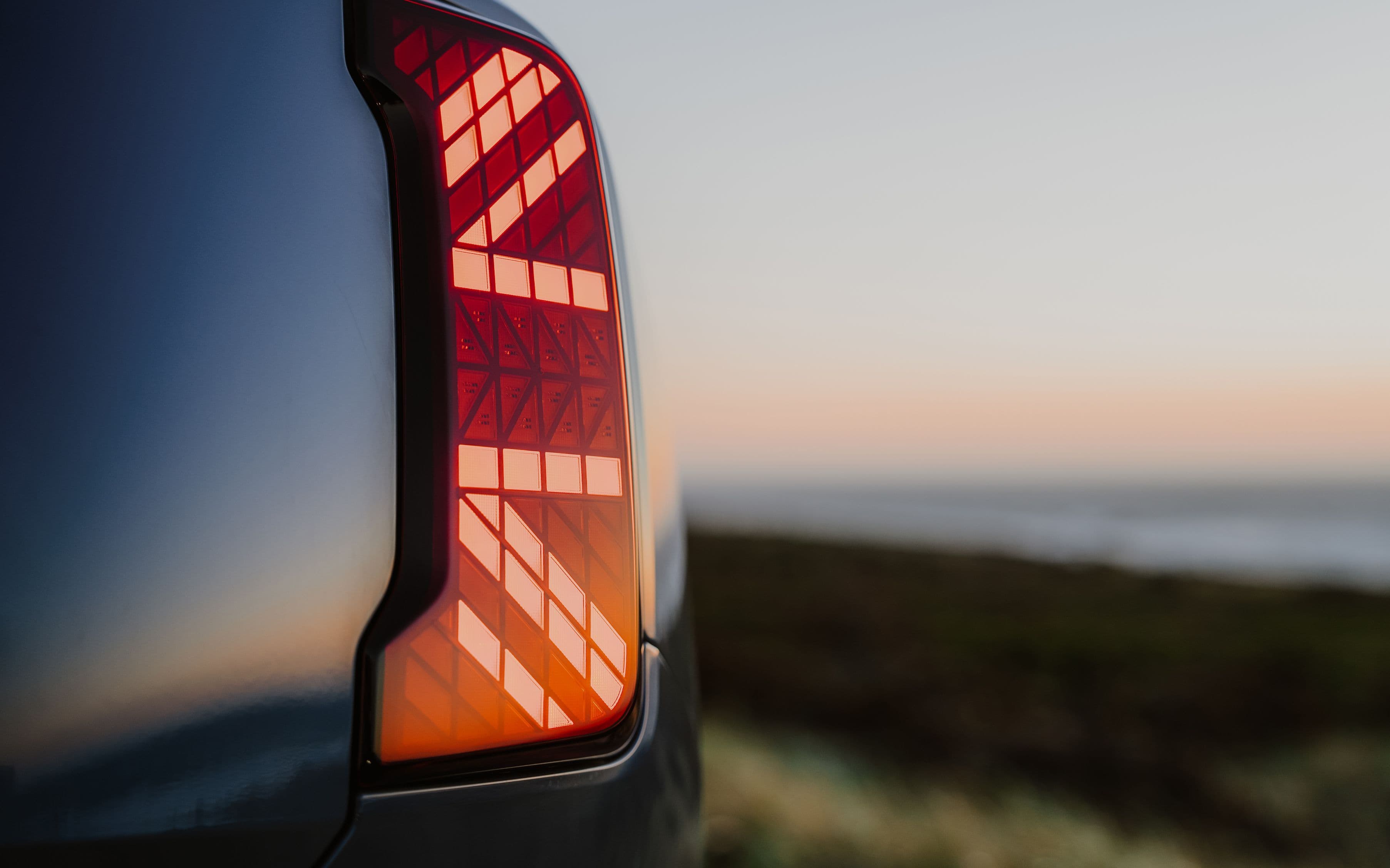 Close-up of a modern car's red LED taillight with a grid pattern illuminated against a dusky sky and ocean background.