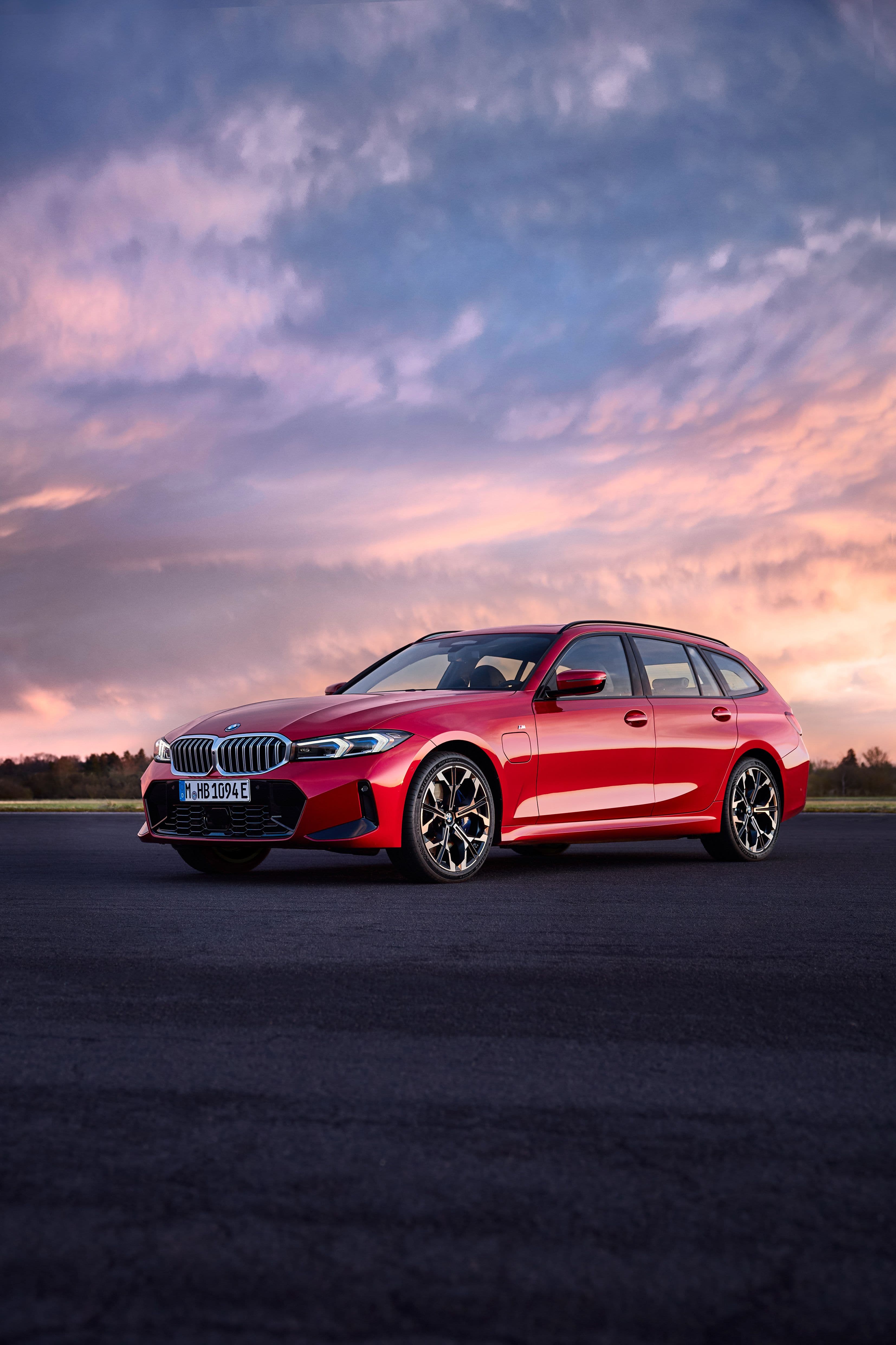 Red BMW 3 Series Touring parked on asphalt road with sunset sky.