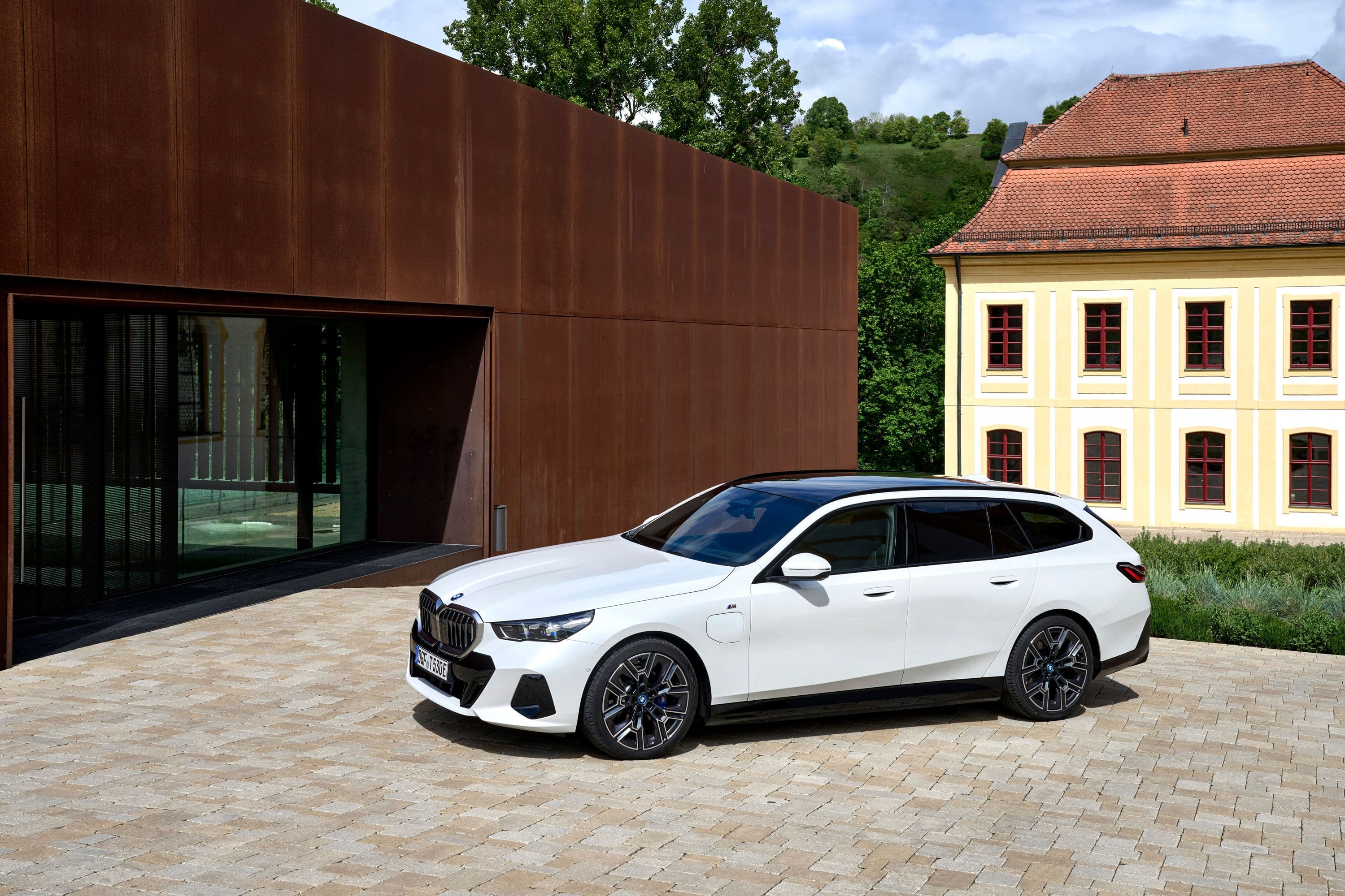 A white BMW 5 Series Touring wagon parked in front of a modern building with a rusted metal facade.