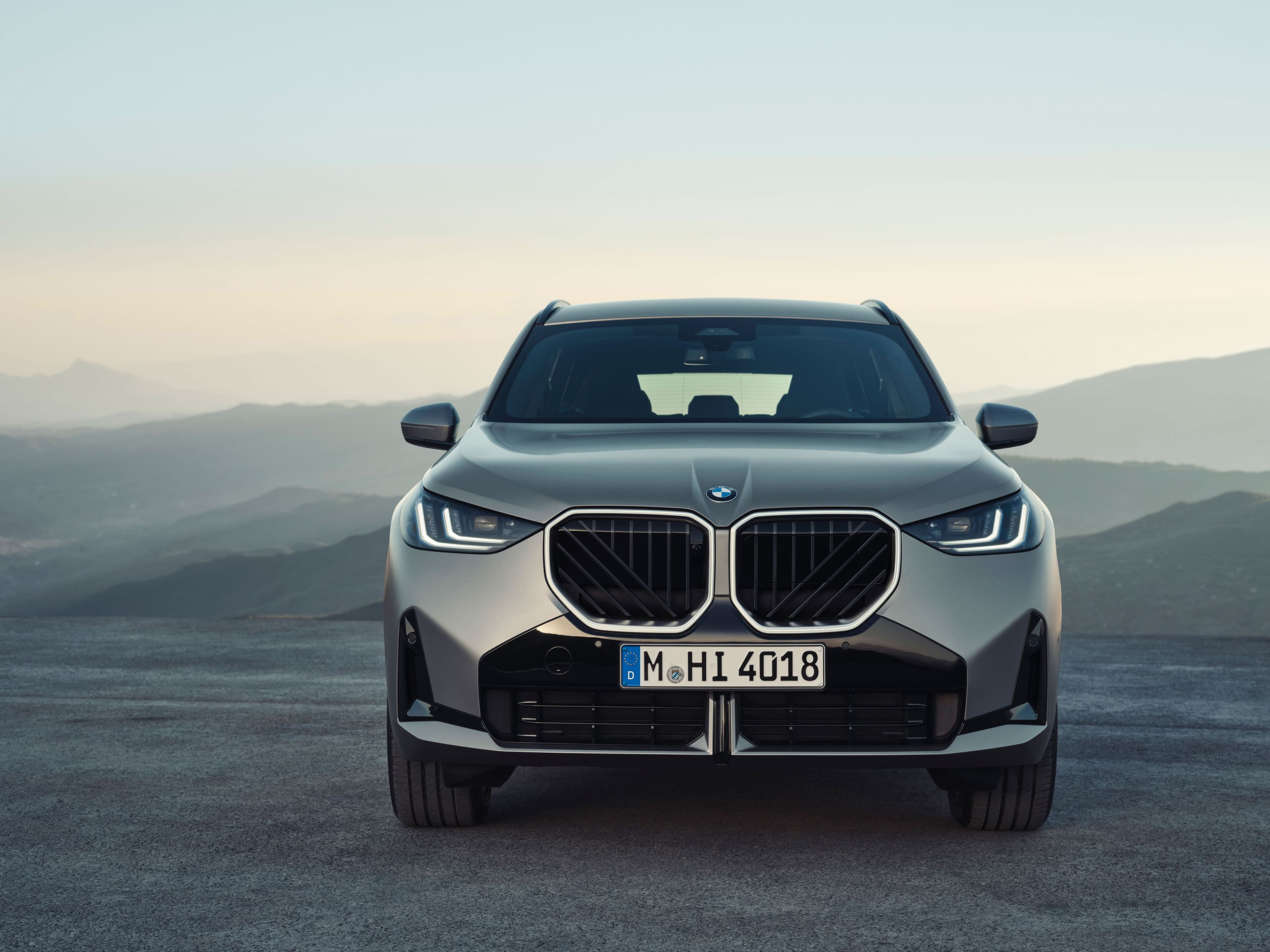 Front view of a silver BMW X1 SUV parked on a gravel road with mountains in the background.