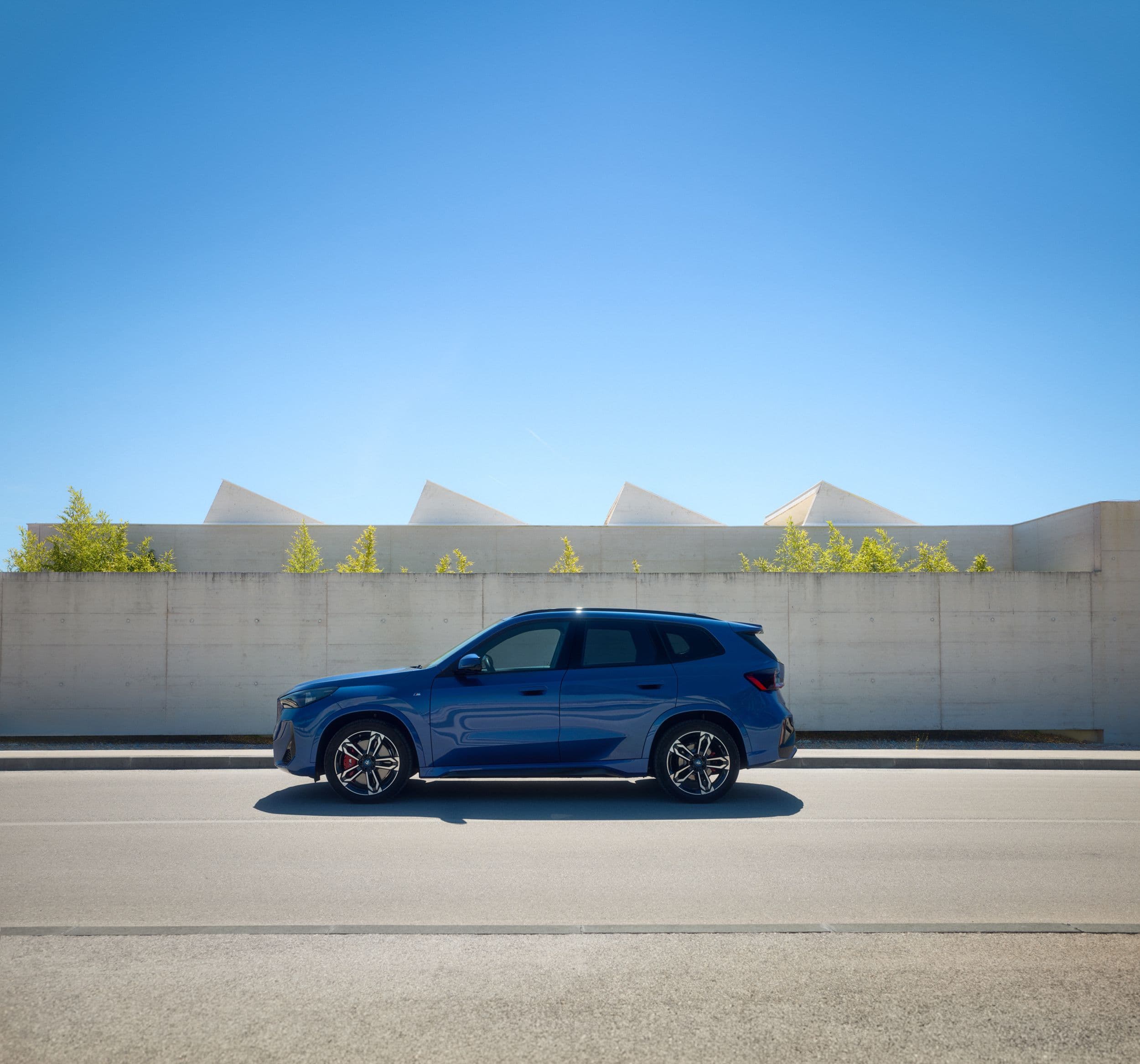 A blue BMW X1 drives down a road with a concrete wall and geometric structures in the background.