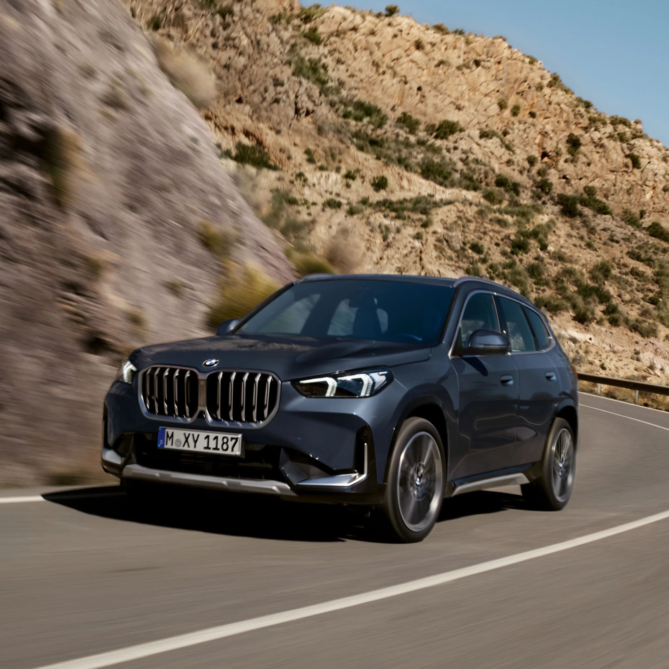 Dark gray BMW X1 drives on a winding road through a mountainous landscape.